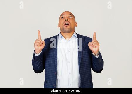 Portrait of astonished young African American businessman guy, amazed and surprised, looking up and pointing with fingers and raised hands. Standing on a gray background Stock Photo