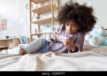 Happy african child girl using mobile apps on smartphone lying on bed at home. Stock Photo