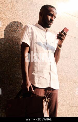 Young serious African businessman in casualwear texting by wall of building Stock Photo