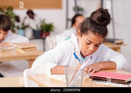 Contemporary mixed-race schoolgirl in whitecoat writing down chemical formula Stock Photo