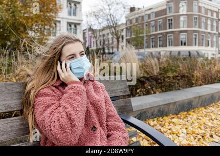 Teenage girl wearing face mask sitting on a bench outside placing a call on her telephone Stock Photo