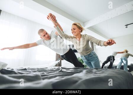 Young happy couple jumping on bed and make fun while sitting at home. Family fool around at and jump on bed Stock Photo