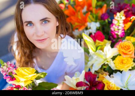 Closeup of beautiful serene young woman with calm expression in a white shirt  surrounded by abundant colorful florist flowers Stock Photo