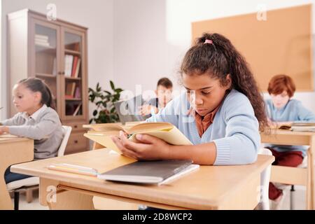Clever cute schoolgirl reading book and making notes by desk in classroom Stock Photo