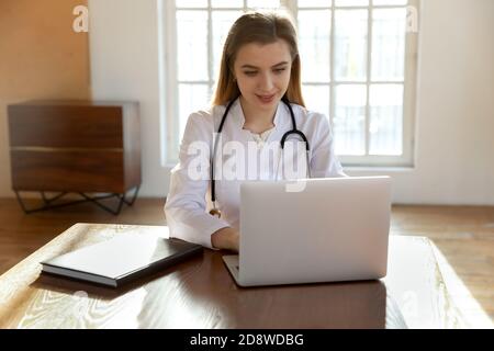 Smiling young female physician sitting at workplace counseling patients online Stock Photo