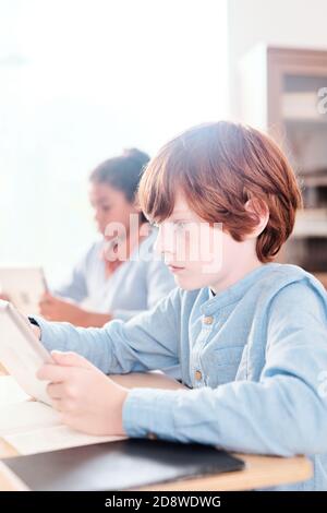 Serious clever schoolkid with tablet looking through text on display at lesson Stock Photo