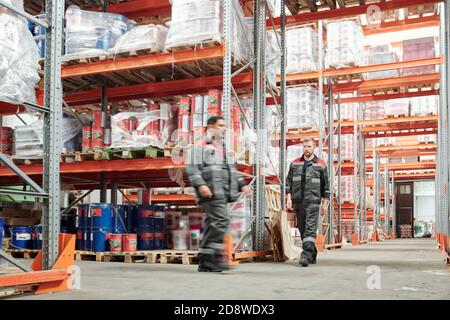 Two young intercultural male workers in uniform moving along huge shelves Stock Photo