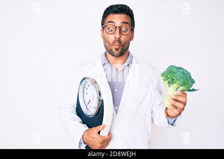 Young hispanic man as nutritionist doctor holding weighing machine and broccoli making fish face with mouth and squinting eyes, crazy and comical. Stock Photo