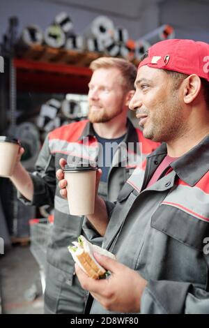 Two happy young warehouse workers having sandwiches and coffee at lunch break Stock Photo