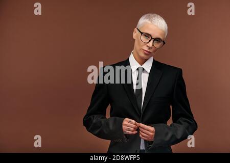 Blond manlike woman in elegant black suit and eyeglasses doing buttons of jacket Stock Photo