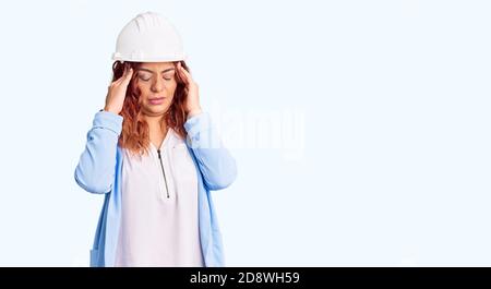 Young latin woman wearing architect hardhat with hand on head for pain in head because stress. suffering migraine. Stock Photo