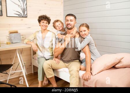 Happy young family of four in casualwear sitting on bed in front of camera Stock Photo