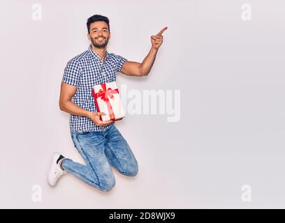 Young handsome hispanic man wearing casual clothes smiling happy. Jumping with smile on face holding birthday gift pointing with finger to the side ov Stock Photo