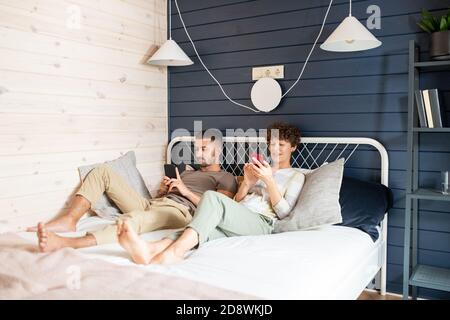 Young restful husband and wife relaxing on bed and scrolling in mobile phones Stock Photo