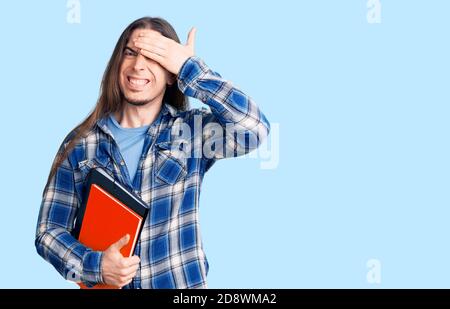 Young adult man with long hair holding collage book stressed and frustrated with hand on head, surprised and angry face Stock Photo