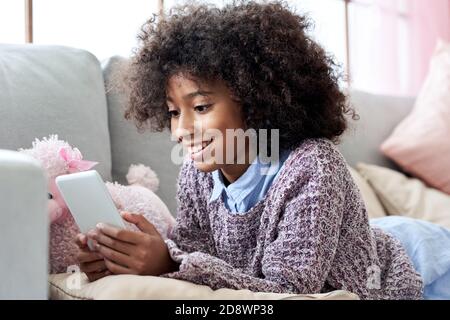 Happy african child girl using mobile apps on phone relaxing on couch at home. Stock Photo