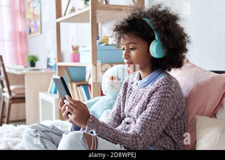 African child girl wearing headphones watching video on phone sitting on bed. Stock Photo