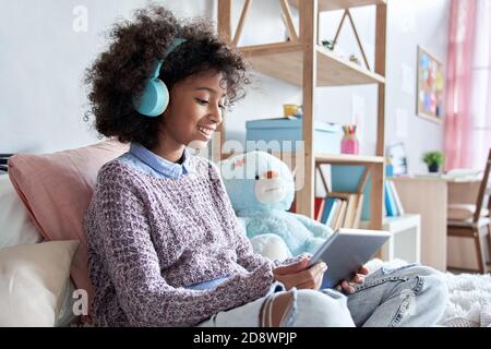 Happy african american kid girl holding digital tablet elearning sitting on bed. Stock Photo