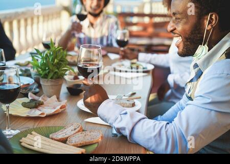 Cheerful multiracial friends eating and drinking wine while wearing protective masks - Focus on black man Stock Photo