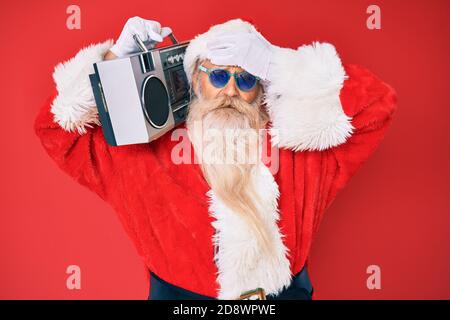 Old senior man wearing santa claus costume and boombox stressed and frustrated with hand on head, surprised and angry face Stock Photo