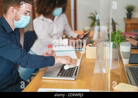 Young worker using sanitizer gel to disinfect his laptop computer inside coworking office - Focus on man eye Stock Photo