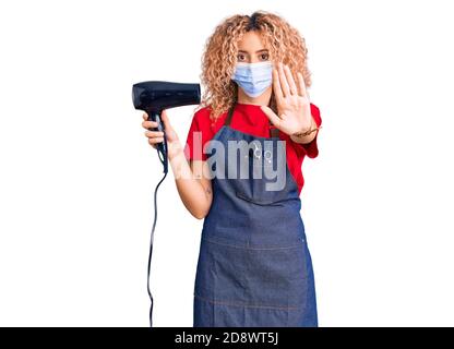 Young blonde woman with curly hair holding dryer blow wearing safety mask for coranvirus with open hand doing stop sign with serious and confident exp Stock Photo