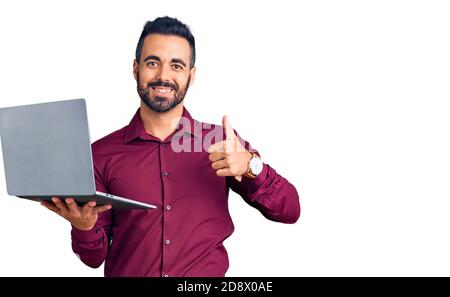 Young hispanic man holding laptop smiling happy and positive, thumb up doing excellent and approval sign Stock Photo