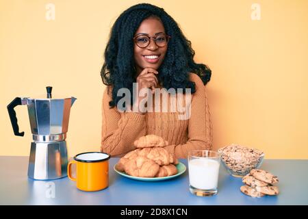 Beautiful african woman sitting on the table eating breakfast in the morning smiling looking confident at the camera with crossed arms and hand on chi Stock Photo