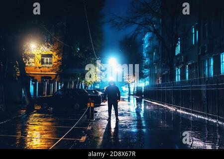 Silhouette of alone stranger in hood at night city street in rain. Creepy killer or stalker, criminal stands in shadow with urban lights reflected in puddles. Thriller horror mysterious atmosphere. Stock Photo
