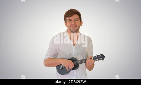 Medium shot. Front view. Young man playing ukulele and signing while walking and looking into camera on gradient background. Professional shot in 4K r Stock Photo