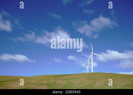 Looking up to white wind turbines against blue sky with light wispy cloud, on grassy knoll. Stock Photo