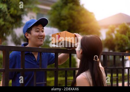 Asian delivery man in blue shirt sending a parcel box to home while a young woman receiving a package in front of custoomer house Stock Photo