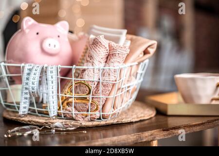 Sewing tools of tailoring scissors, tape measure, pink lace fabric in mesh basket and thread spools and piggy bank. Stock Photo