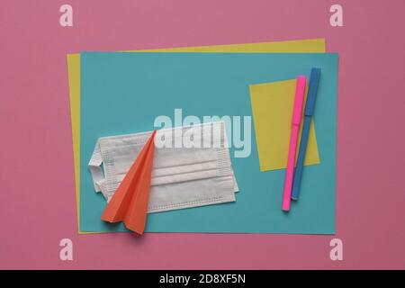 Face mask on top of coloring pens and papers with paper plane. Pink background. Stock Photo