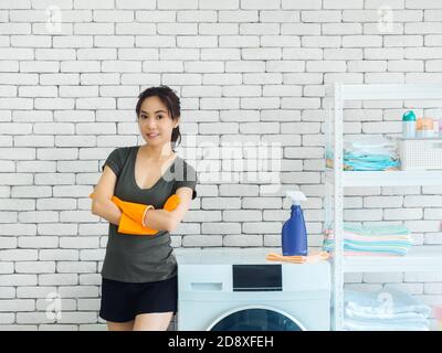 Beautiful smiling Asian woman, happy housewife wearing orange protective rubber gloves standing with arms folded beside washing machine on white brick Stock Photo