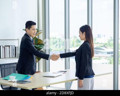 Asian businessman in suit and businesswoman shaking hands in office. Smiling manager standing at his desk in his office handshaking with young female, Stock Photo