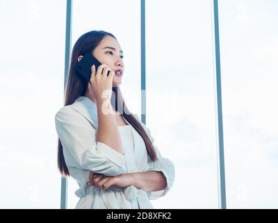 Young pretty Asian woman wearing formal white shirt talking on the phone call near huge glass window in office with copy space. Portrait of beautiful Stock Photo