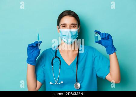 Concept of covid-19 and quarantine concept. Cheerful smiling female doctor in medical mask, latex gloves and scrubs, showing syringe filled with Stock Photo