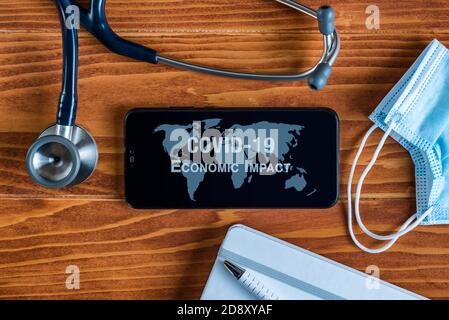 Coronavirus COVID-19 concept: a smartphone sorrounded by a stethoscope, a medical mask and other objects with a world map and an Economic Impact text Stock Photo