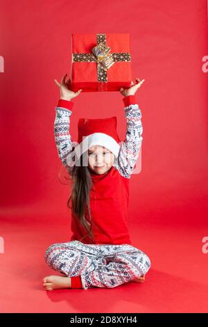 smiling girl in pajamas and a Santa Claus hat, barefoot sitting on the floor holding a present over his head, isolated Stock Photo