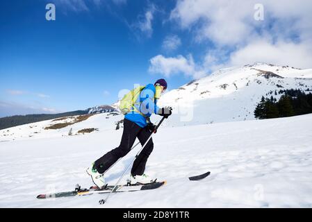 Happy tourist in colorful clothing and sunglasses with backpack climbing on skis in deep snow on background of bright blue sky and beautiful mountain. Winter vacations, active lifestyle concept. Stock Photo