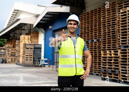 Portrait of young Indian worker working in logistic industry outdoor in front of factory warehouse. Smiling happy man with hard hat looking at camera giving thumbs up gesture at depot Stock Photo