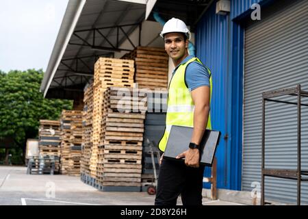 Portrait of young Indian worker working in logistic industry outdoor in front of factory warehouse. Smiling happy man with hard hat looking at camera walking and holding a laptop at depot Stock Photo