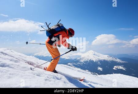 Sportsman skier in skiing equipment with backpack riding down steep slope in deep snow on copy space background of blue sky and beautiful mountain landscape. Winter sports, courage and speed concept. Stock Photo