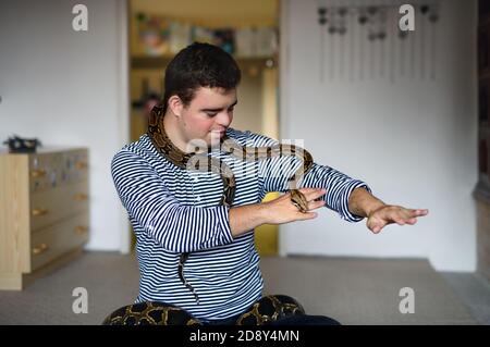 Down syndrome adult man sitting indoors in bedroom at home, playing with pet snake. Stock Photo
