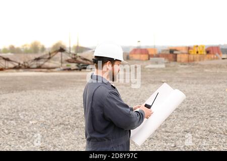 Back view of building engineer standing on construction site, talking by VHF walkie talkie and holding blueprints. Stock Photo