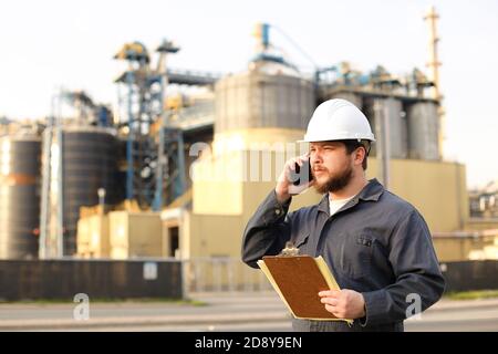 Industrial worker talking by VHF walkie talkie and holding papers near factory outside. Stock Photo