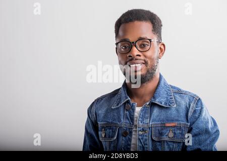 Horizontal close up portrait of joyful happy african guy in spectacles denim jacket smiling broadly with teeth laughing looking at camera over gray ba Stock Photo