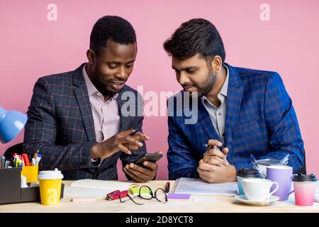 Two confident serious business people wearing suits sitting at desk in office. Dark skinned manager holding smartphone and looking at screen of gadget Stock Photo