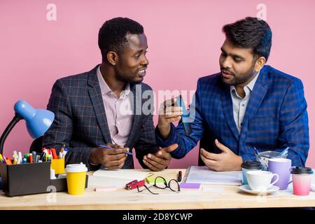 Two diverse business people in suits chatting, sitting at table in office. African young man showing his colleague new app on his phone, using mobile Stock Photo